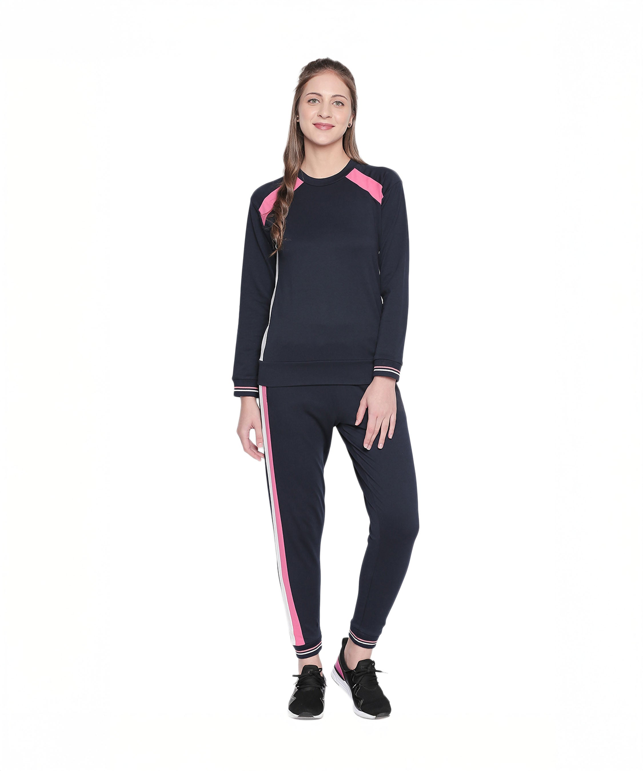 Woman in a full sleeve Navy Blue round neck sweatshirt and pink tracksuit, paired with black sneakers, stands smiling against a white background.