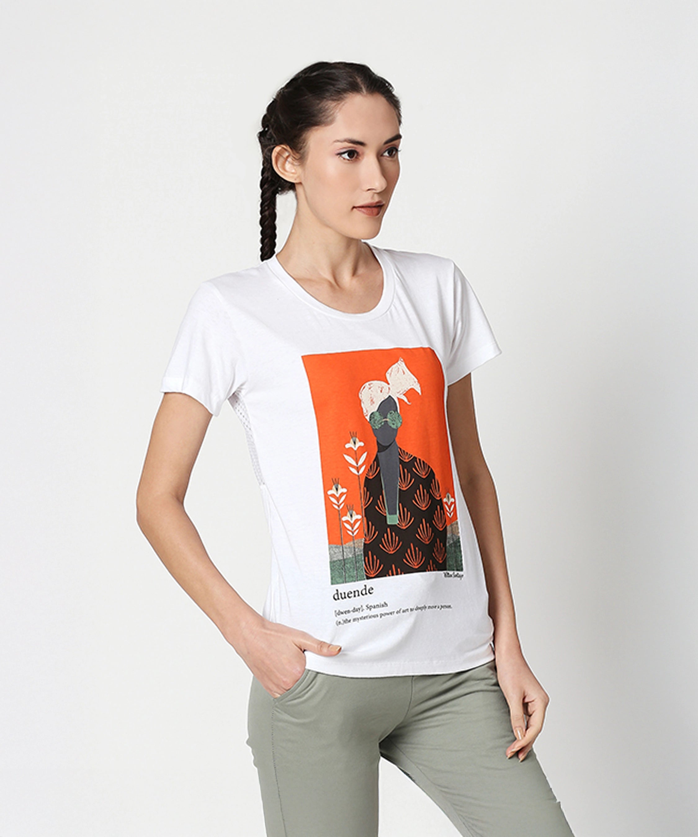 A woman wears the Duende Printed cotton tee—an artistic graphic T-shirt made from breathable cotton—and green pants, posing with one hand in her pocket against a plain background.