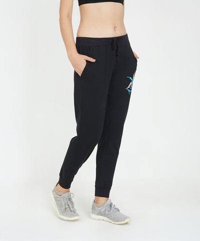 Woman wearing Ashtanga Series Black Joggers, a black sports bra, and gray sneakers, standing with hands in pockets.