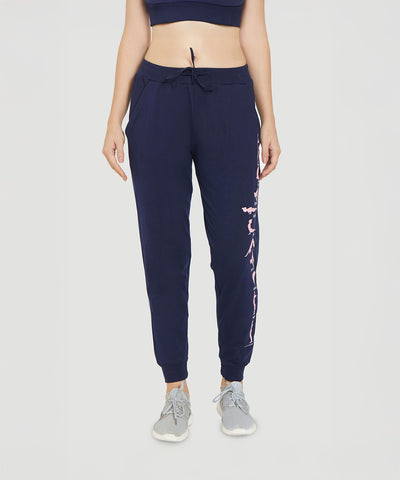 Woman wearing Ashtanga Series Blue joggers featuring a pink floral graphic on the left leg, styled with grey sneakers and a navy crop top.