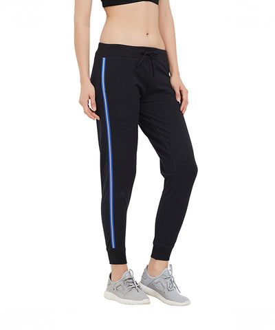A woman stands wearing Classic Joggers TLC02SS in blue with sporty side stripes and a tapered fit, paired with gray sneakers and a black sports bra.