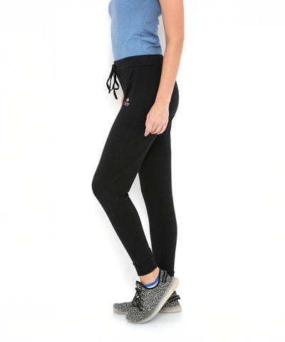 Woman in blue top and Black Classic Joggers TLC02 stands sideways in black and white sneakers. The joggers offer a slim tapered fit for easy, stylish comfort, shown against a crisp white background.