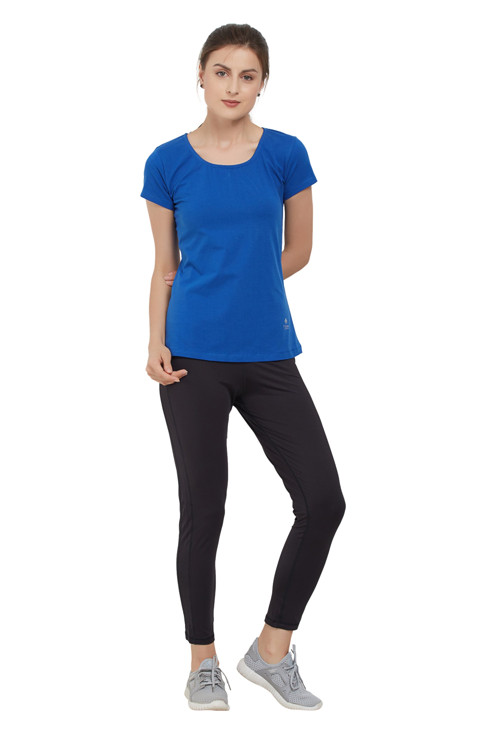 A woman in a Royal Blue Round Neck Tee and black leggings stands with one hand behind her back, wearing moisture-wicking grey sneakers.