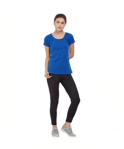 A woman in a TUNA LONDON Royal Blue Round Neck Tee and black pants stands with one arm behind her back against a white background. The cotton lycra tee offers comfort and style.