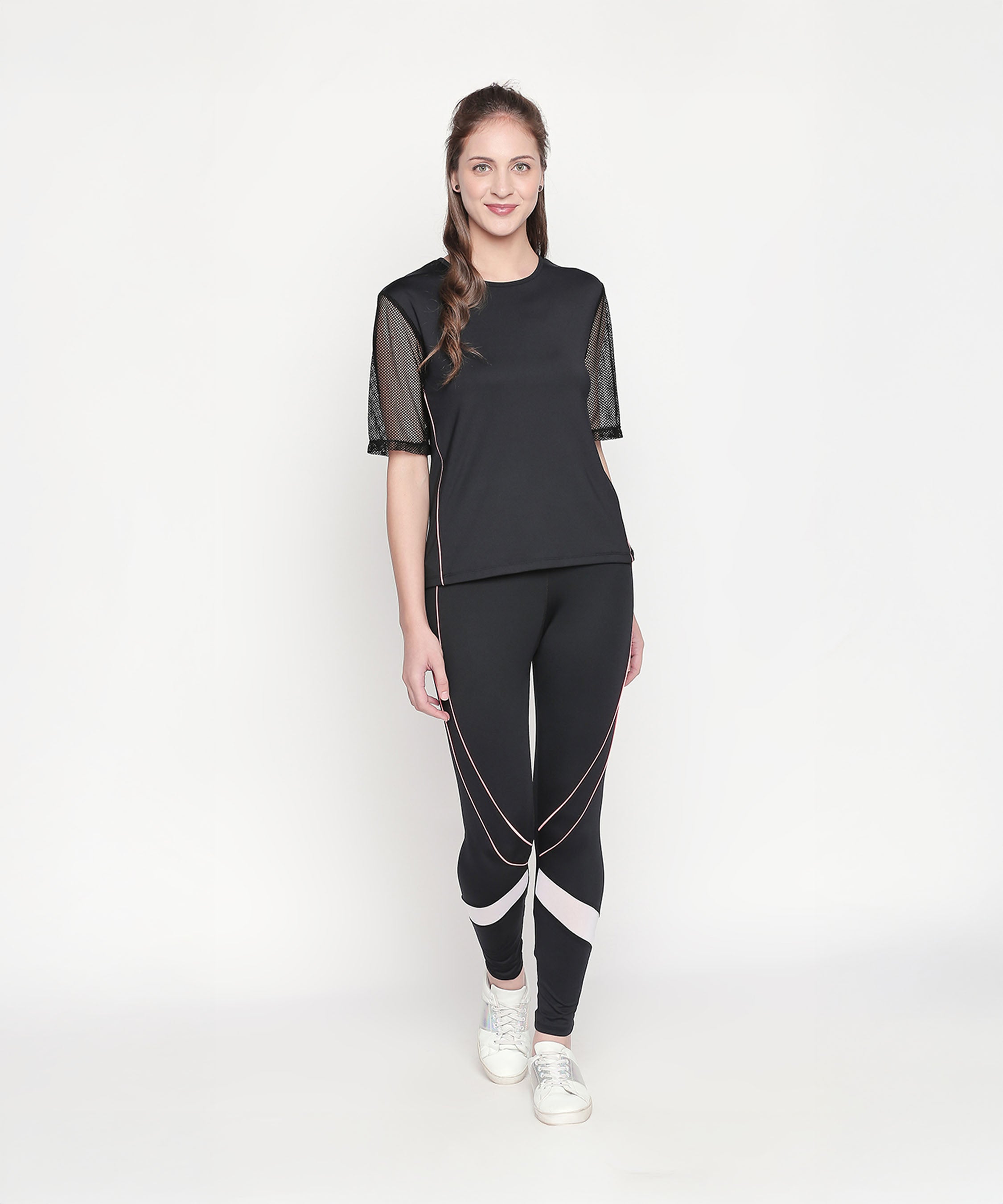 Woman wearing the Black Loose Fit Round Neck Net Half Sleeve Top, styled with white sneakers. The top’s moisture-wicking fabric keeps you cool during any workout. She stands against a white background.