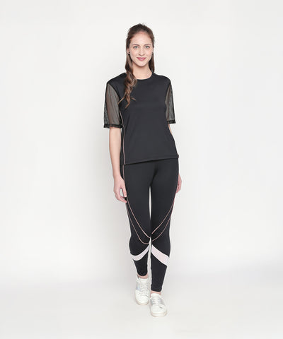 Woman wearing the Black Loose Fit Round Neck Net Half Sleeve Top, styled with white sneakers. The top’s moisture-wicking fabric keeps you cool during any workout. She stands against a white background.
