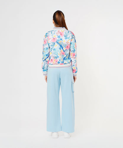 A woman faces away on a plain white background, wearing the Blossom Blue Set with Crop Top featuring light blue cargo track pants and a floral bomber jacket.