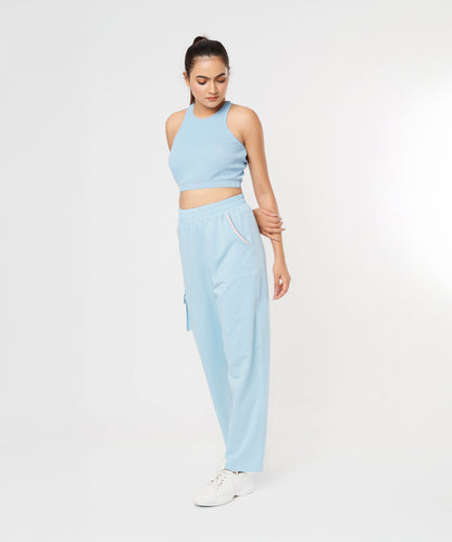 Woman wearing the Blossom Blue Set with a light blue sleeveless crop top and cargo track pants, standing and looking down against a white background.