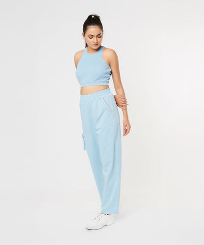 Woman wears the Blue Dream Crop Top—a powder blue racerback athleisure piece—with matching joggers and white sneakers, standing while looking down.