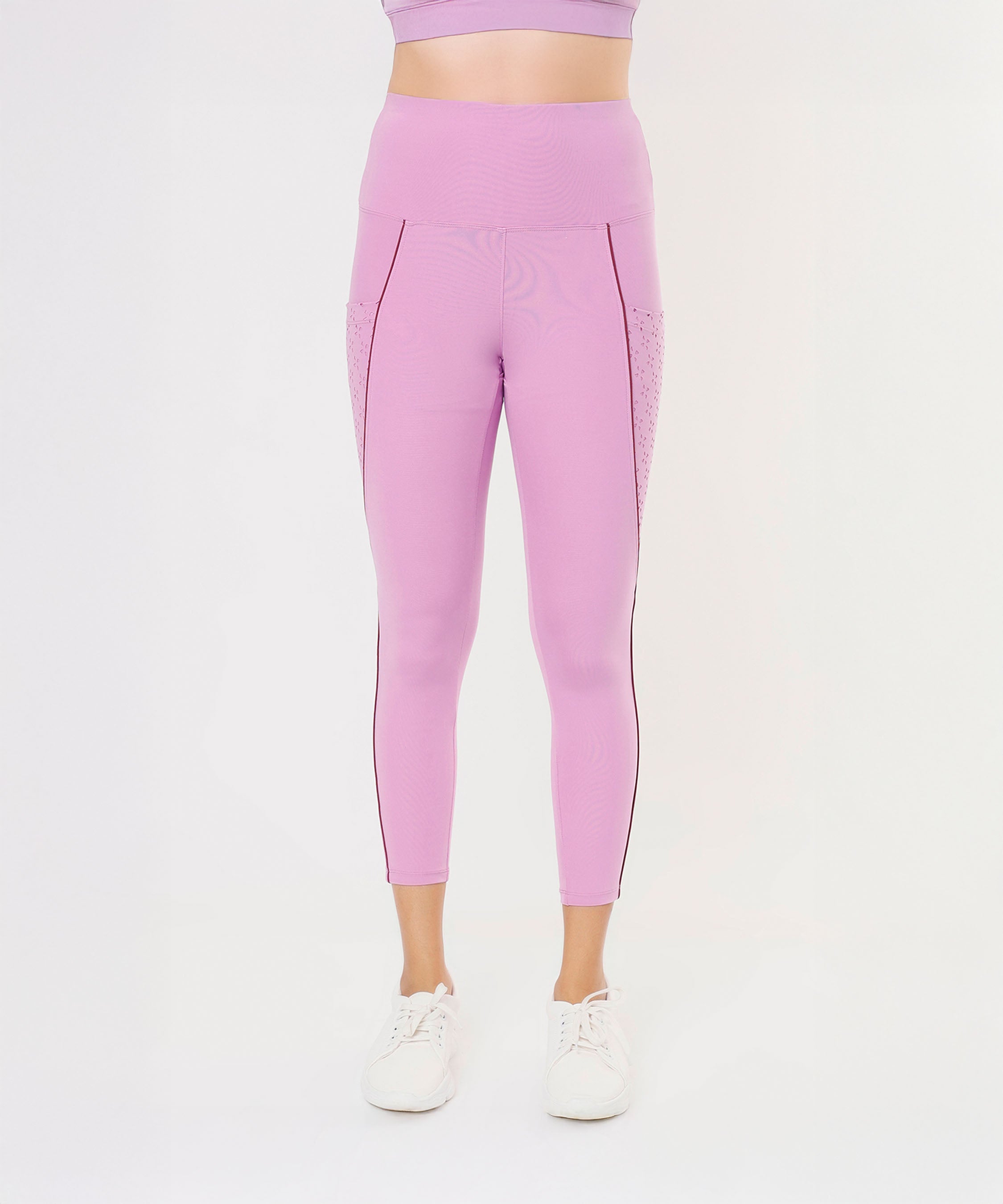 A woman stands against a white background wearing Tuna Active Pink Lilac Leggings—high-waisted with a laser cut pocket—paired with white sneakers.