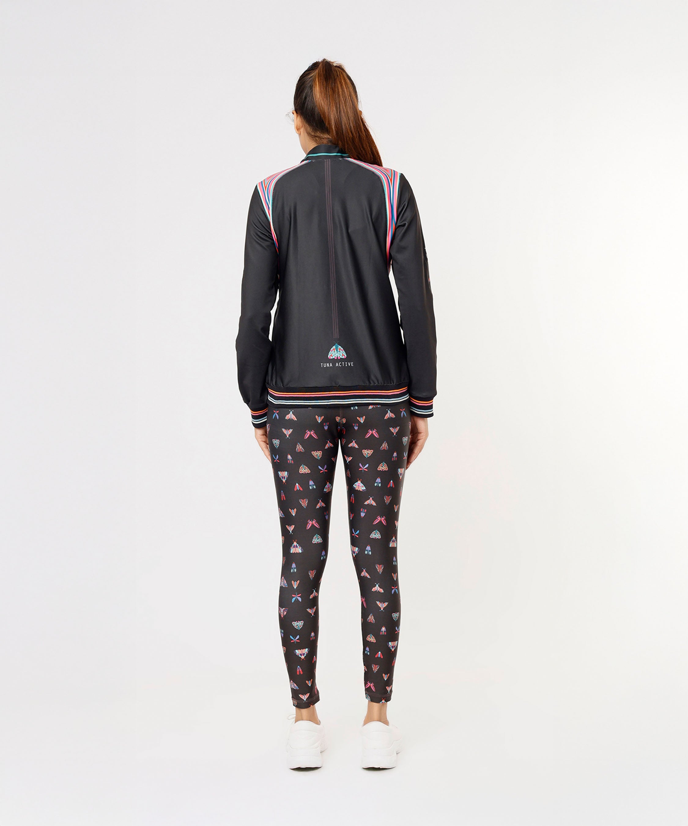 Woman standing facing away, wearing the Tuna Active Black Bomber Jacket with patterned leggings and white sneakers.