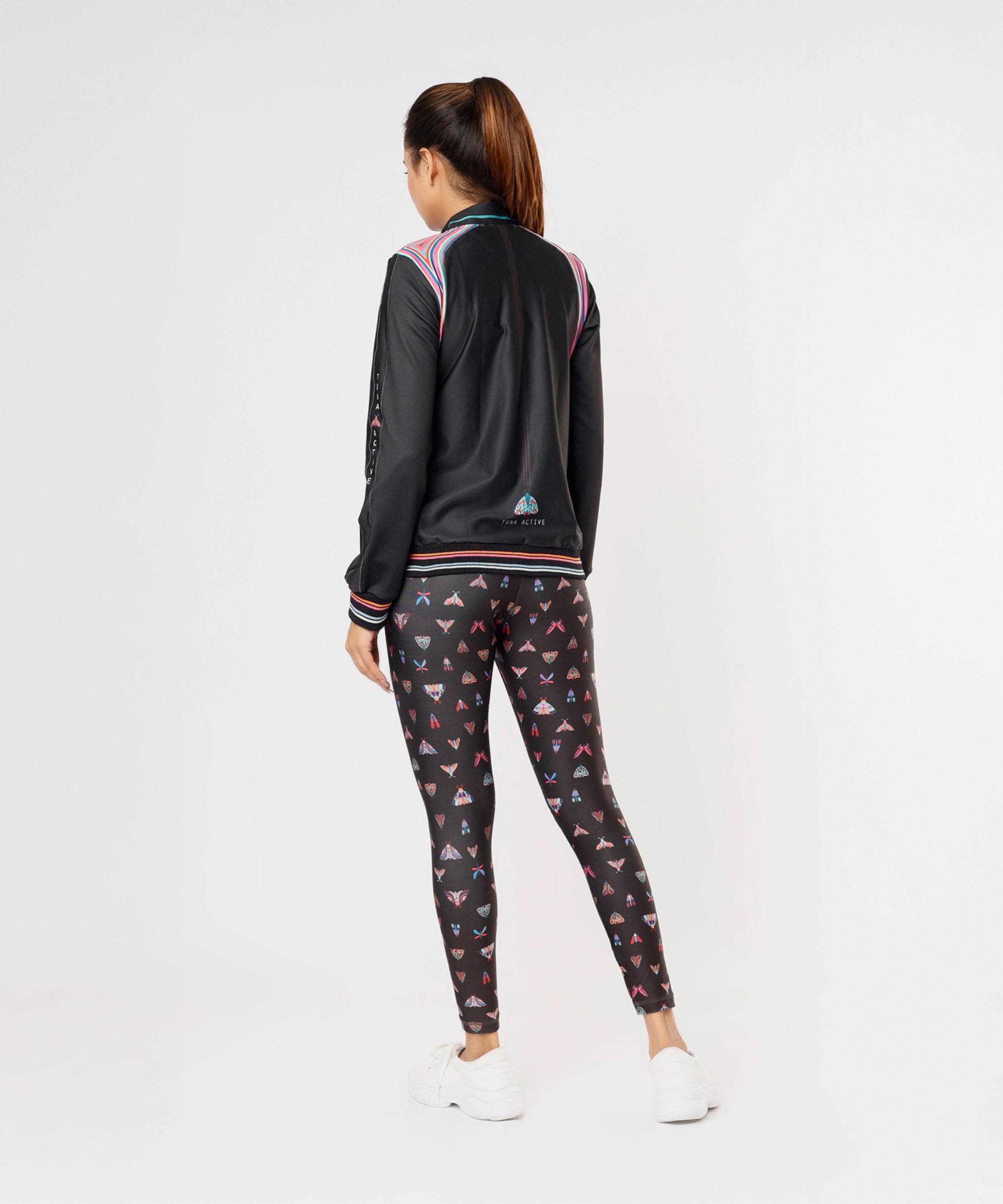 Woman standing with her back to the camera, wearing a Tuna Active Black Bomber Jacket, butterfly print leggings, and white sneakers.