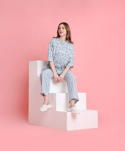 A woman in relaxed-fit Linen Blue loungewear sits on white steps with a pink background, smiling softly.