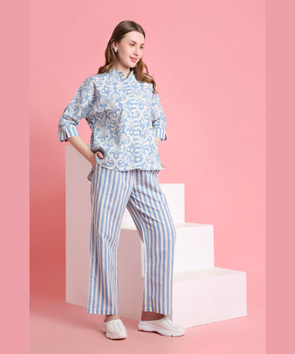 Woman in a Linen Blue damask-print top and striped pants poses by white stairs against a pink background.