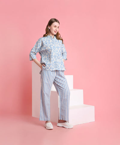 A woman in a relaxed-fit Linen Blue patterned top and striped pants stands near white steps against a pink background.