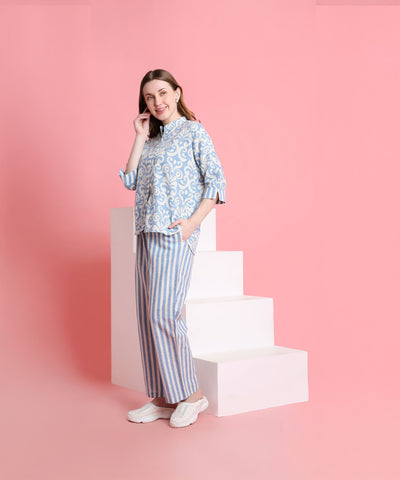 Woman in Linen Blue pyjama set stands by white steps against a pink background, smiling.