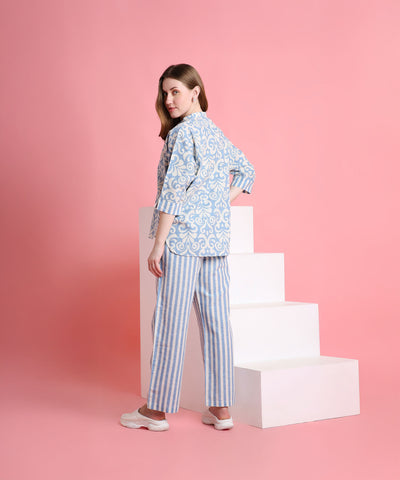 A woman in the Linen Blue set stands by white stairs against a pink background, looking over her shoulder.