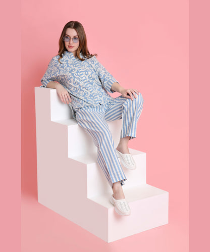 A woman in Linen Blue damask-print top and relaxed-fit pyjamas sits on white steps with a pink background, wearing sunglasses.