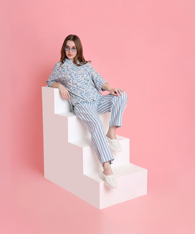 Woman in the Linen Blue pyjama set sits on white steps against a pink background, pairing her bold look with sunglasses.