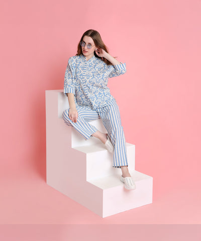 A woman in the relaxed-fit Linen Blue pyjama set and sunglasses sits on white geometric steps with a pink backdrop.