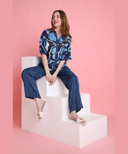 Smiling woman in the Ocean Breeze blue patterned shirt and striped pants, made from breathable fabric, sits on white steps with a pink background.