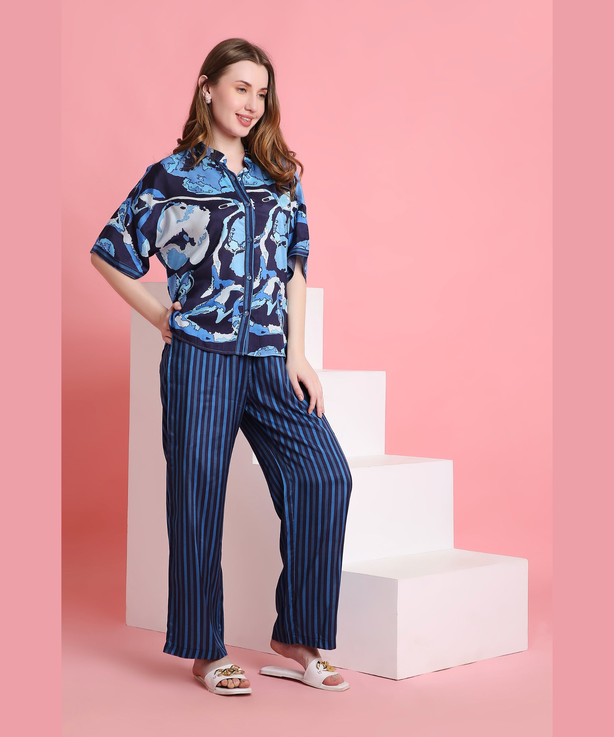 A woman wears the Ocean Breeze outfit—a blue patterned shirt and striped pants made from breathable fabric—while standing by white steps against a pink background.