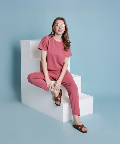A woman wearing a Vintage Pink Knit Cord Set and sandals sits on white steps against a blue background, smiling at the camera.