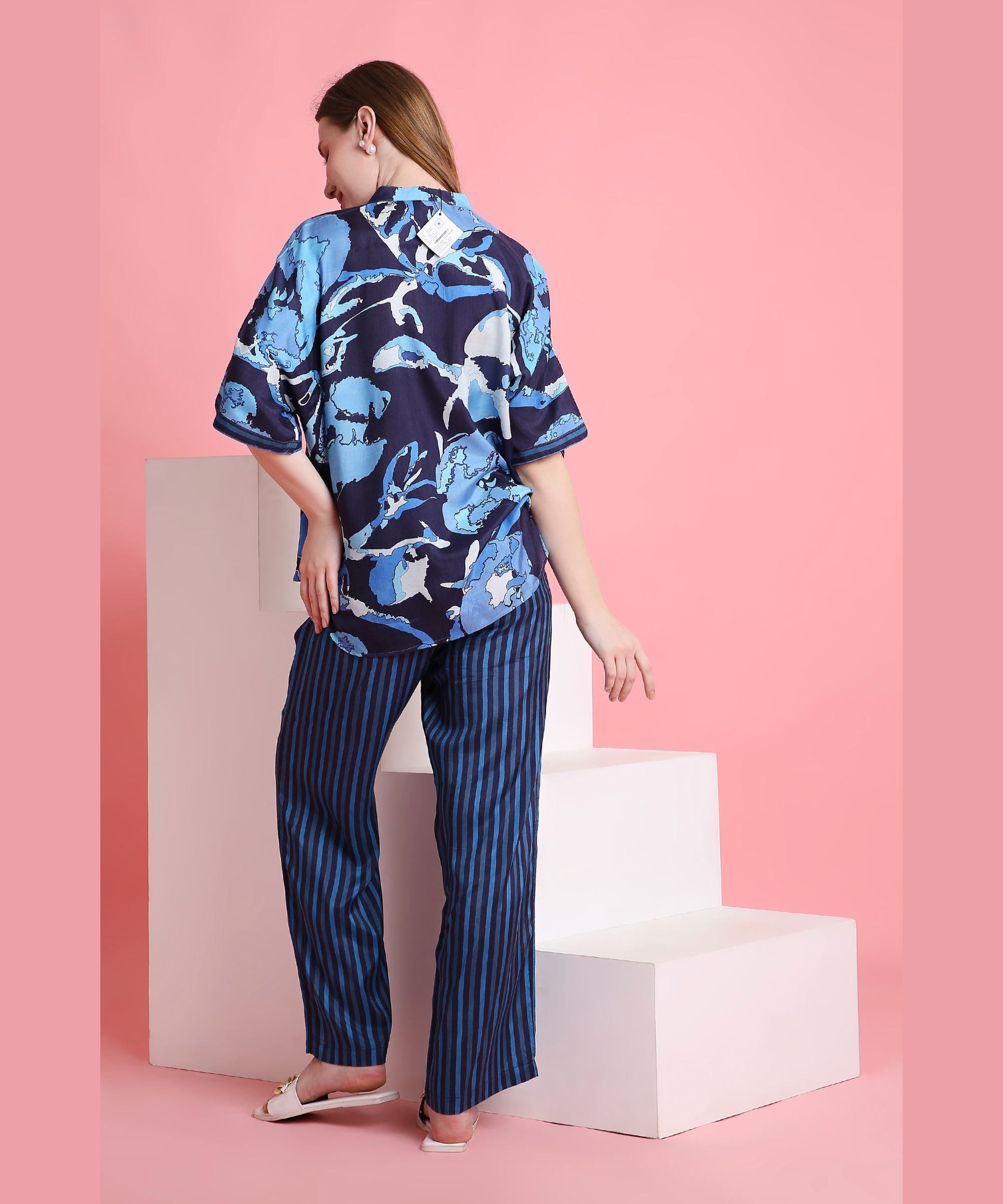 A woman poses with her back turned in Ocean Breeze—a blue floral shirt and striped, breathable pants—set against a pink background.