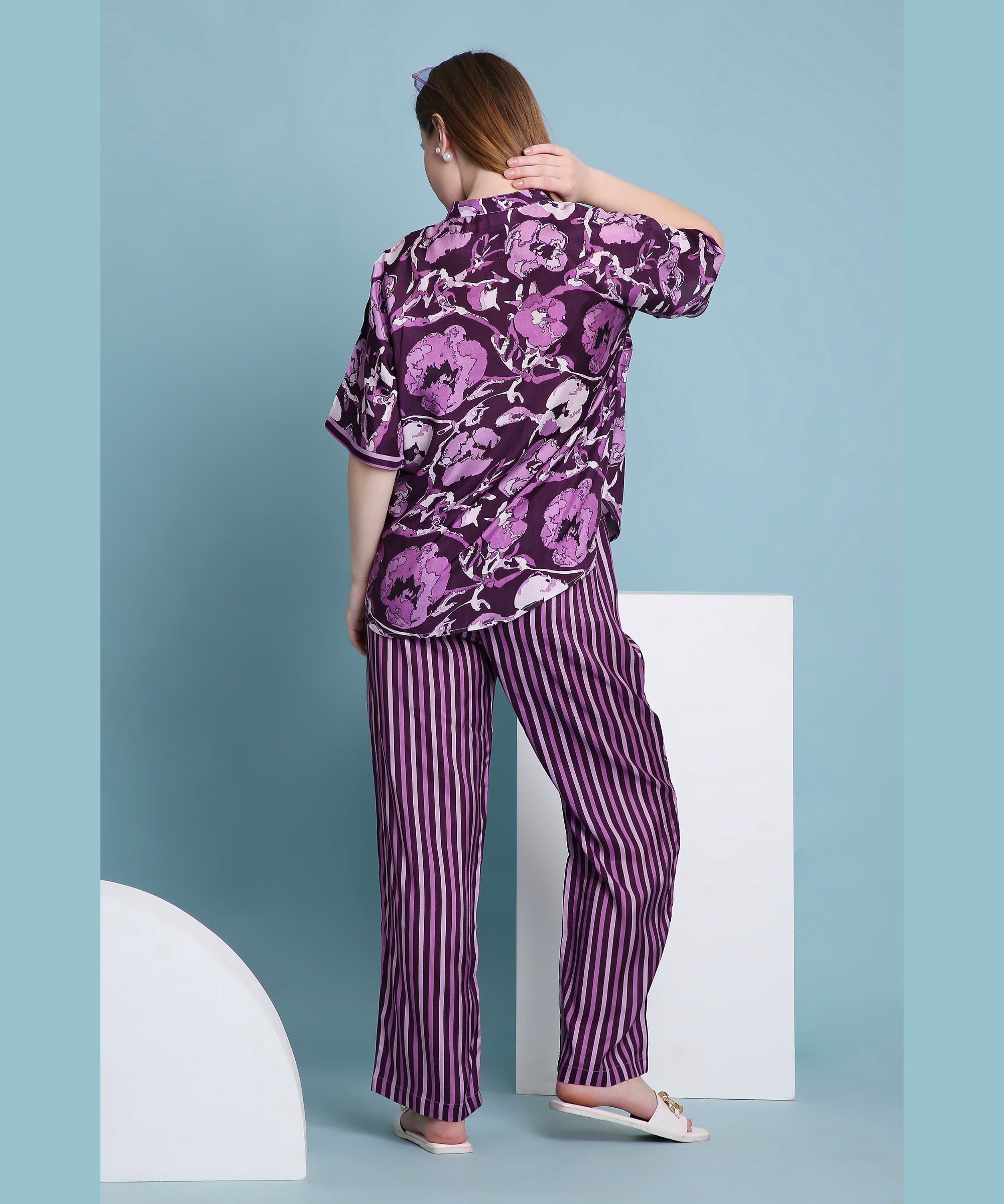 A woman models the Purple Breeze pyjama set, featuring floral and striped designs, as she stands facing away against a blue background.