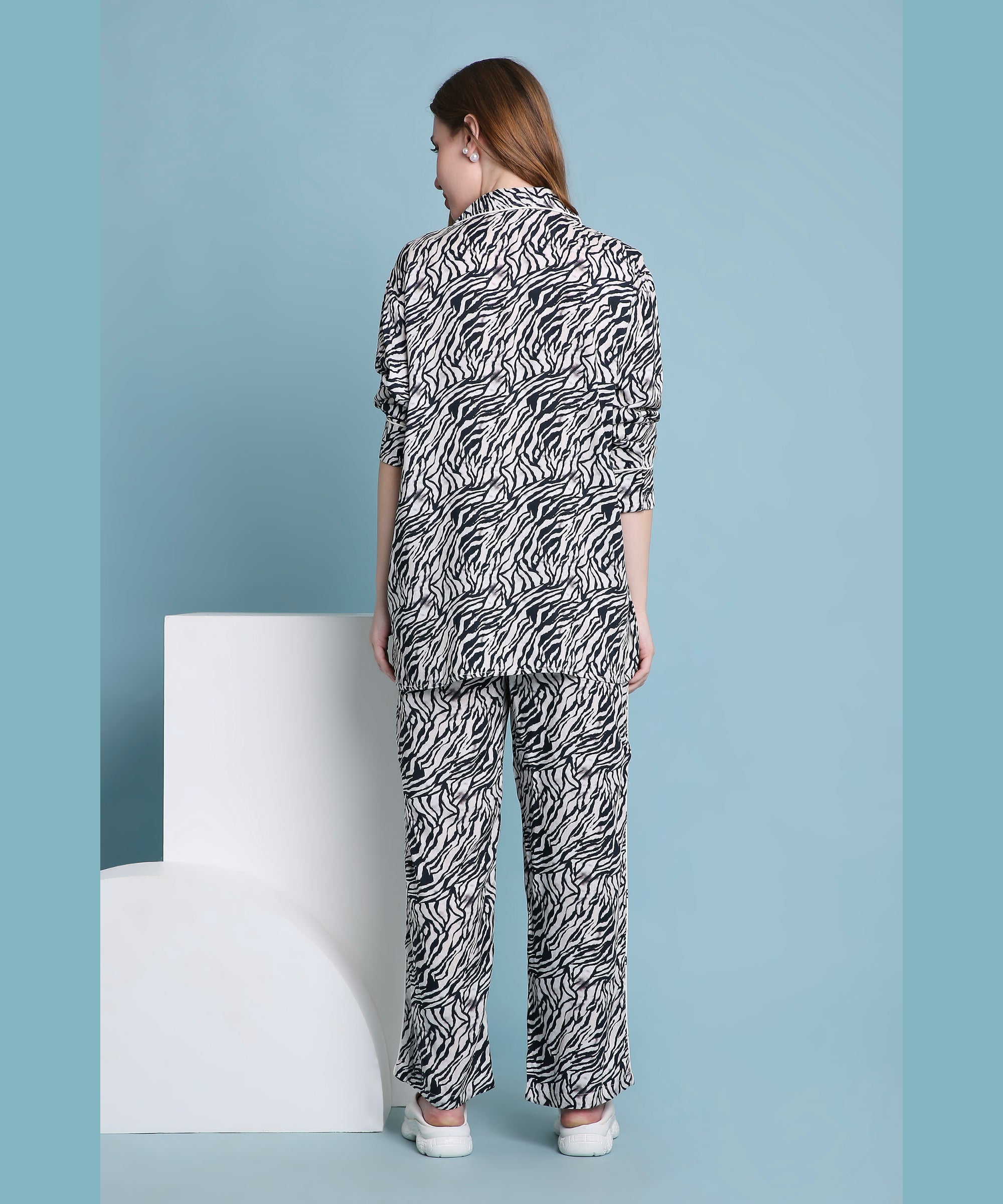 A woman wears the Zeba relaxed fit pyjama set featuring a zebra print, standing with her back turned against a light blue background with geometric white shapes.