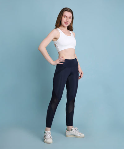 A woman smiles at the camera against a plain blue background, wearing indigo poly blended leggings and a white sports bra.