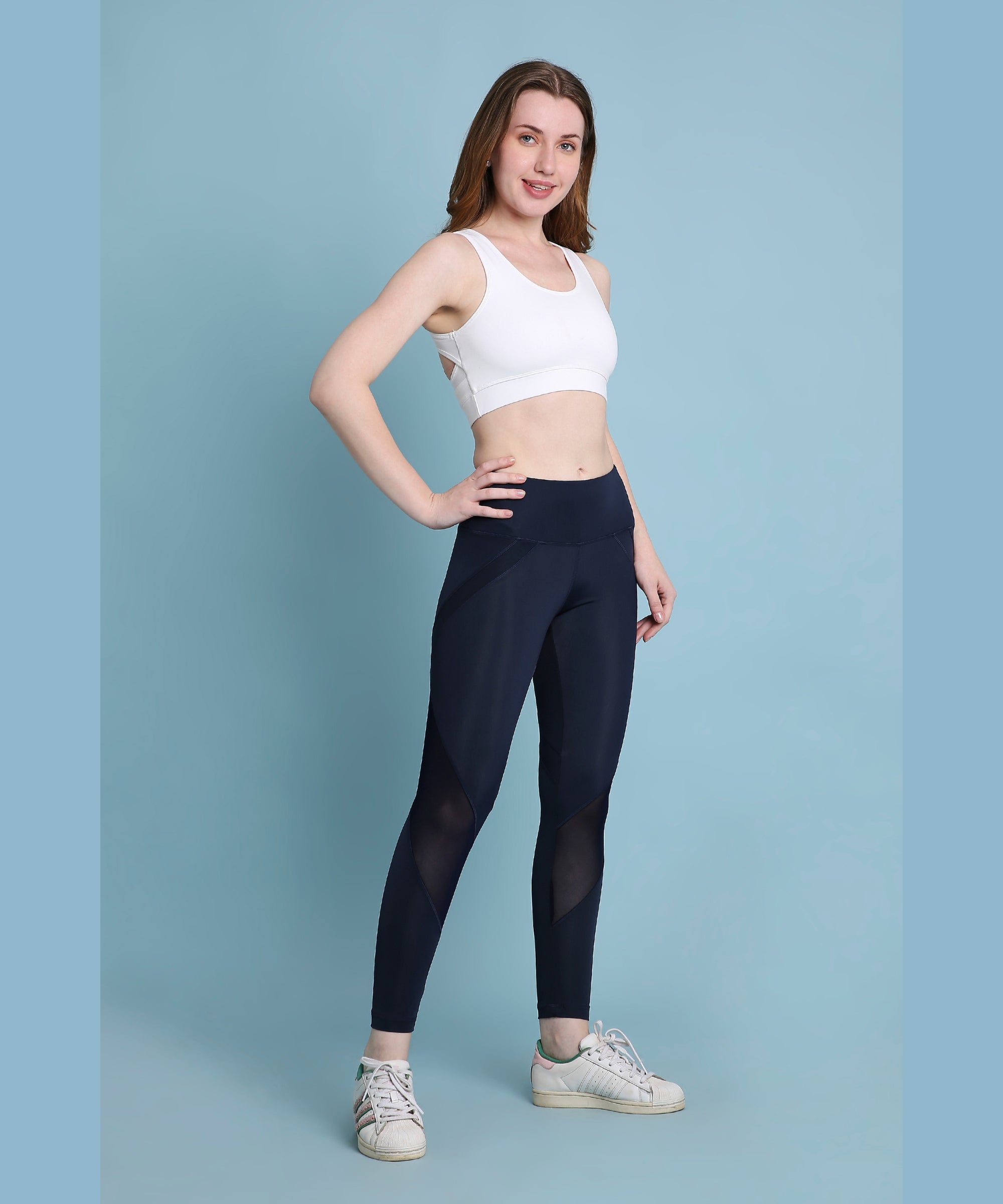 A woman smiles with one hand on her hip, posing against a blue background while wearing indigo poly blended leggings and a white sports bra.