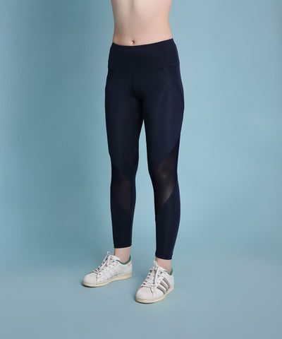 A person wearing Indigo poly blended leggings and white sneakers stands against a plain blue background, with their torso not visible.