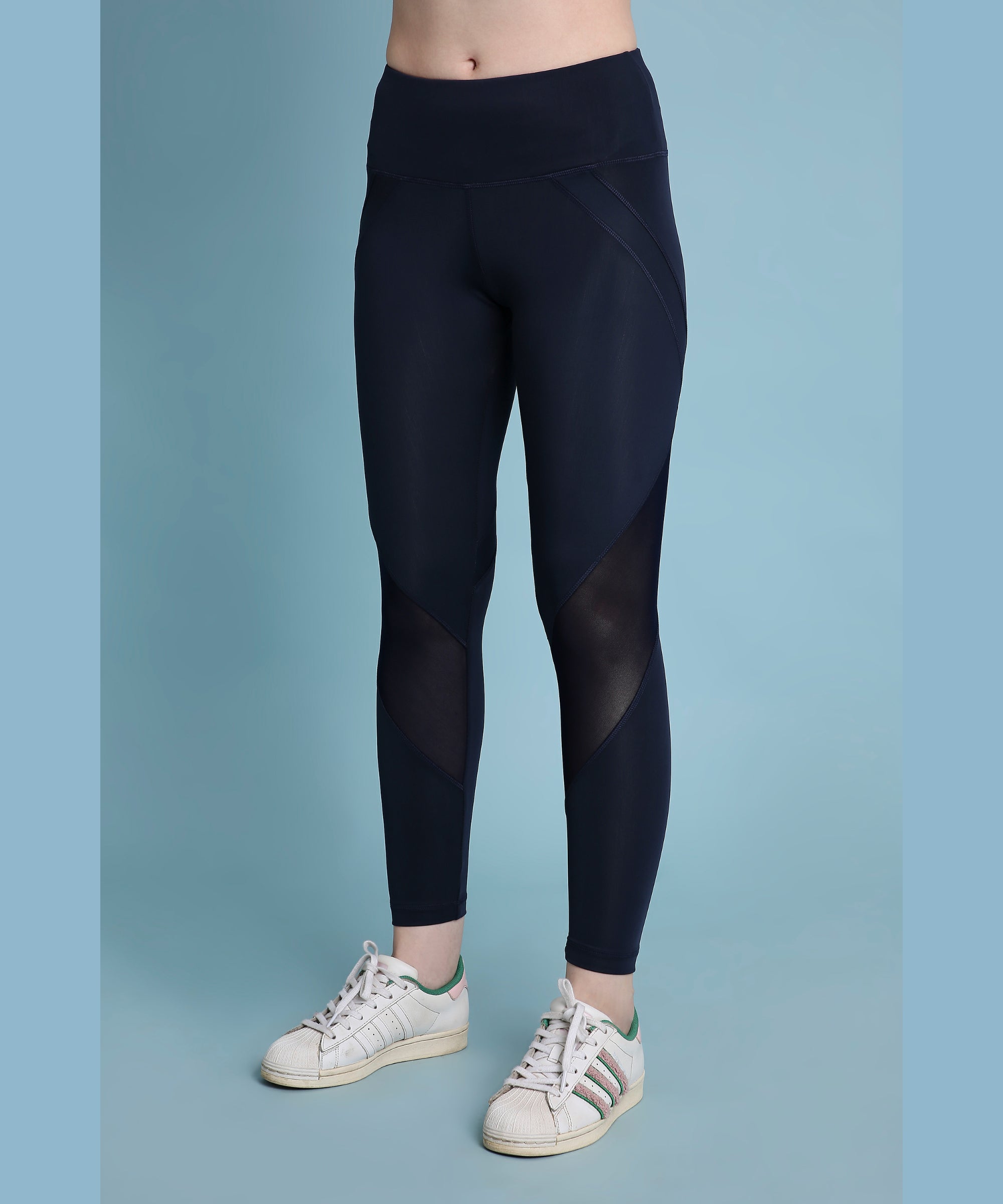 A person wearing Indigo poly blended leggings and white sneakers stands against a blue background.