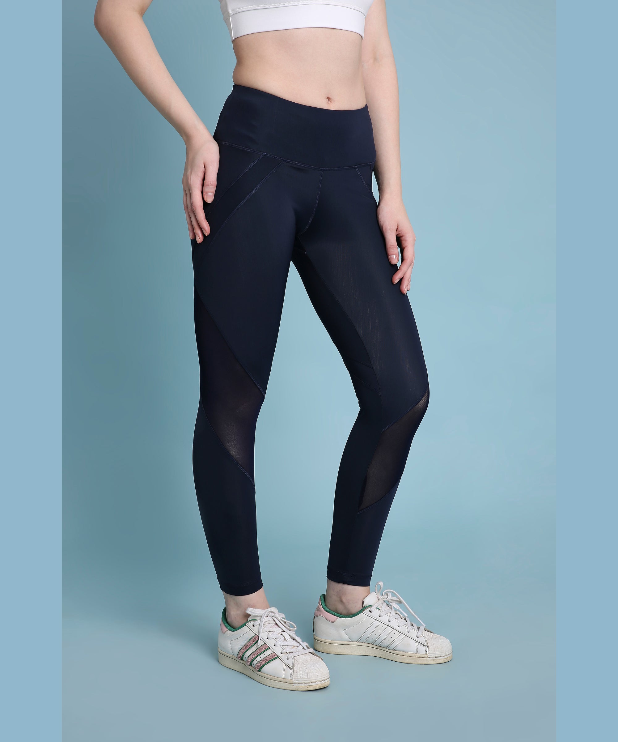 A woman stands confidently against a blue background wearing indigo poly blended leggings, white sneakers, and a white crop top.