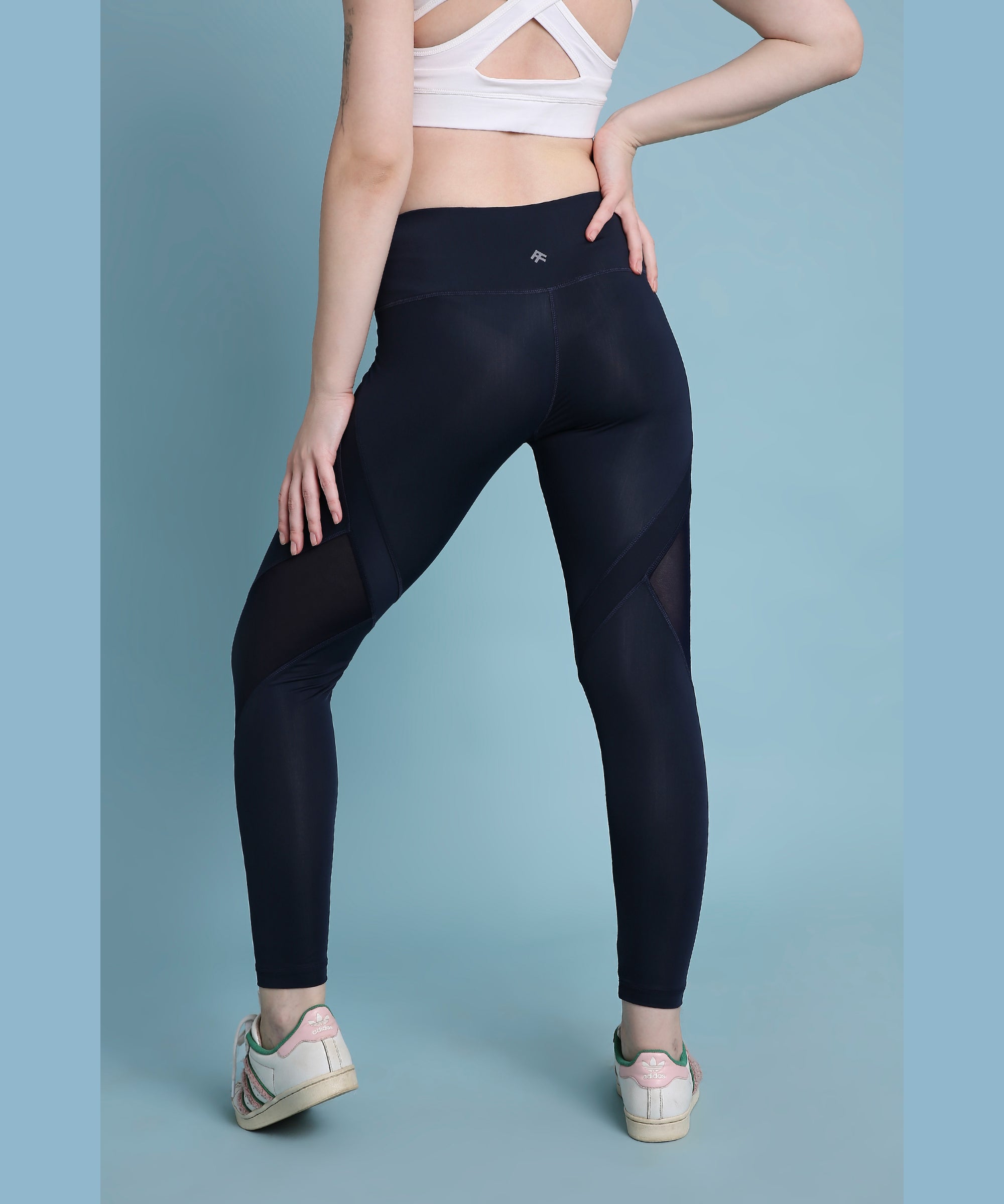 A person wearing Indigo poly blended leggings and a white sports bra stands against a blue background, facing away.