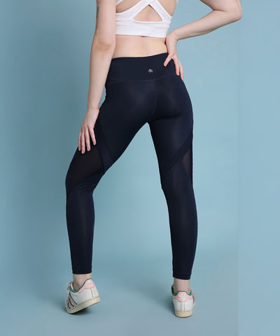 A woman stands facing away against a light blue background, wearing indigo poly blended leggings and a white sports bra.