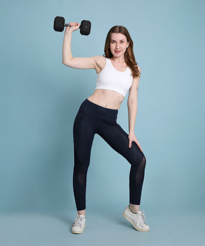 A woman in athletic wear holds a dumbbell above her head against a light blue background, highlighting her stylish and breathable Indigo poly blended leggings.