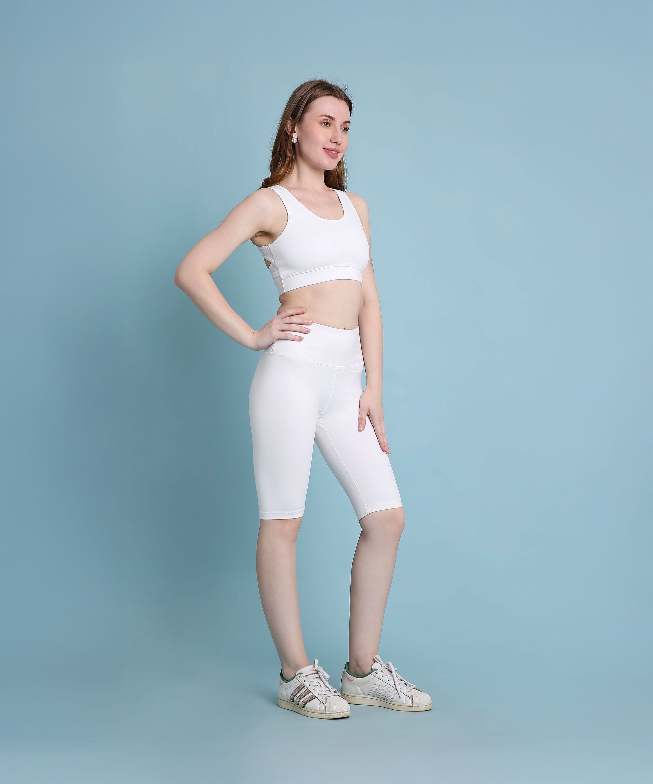 A woman in white sportswear and sneakers poses confidently against a light blue background, showcasing the White Sports Bra, which offers medium support and is made from moisture-wicking fabric.