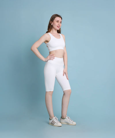 A woman in white sportswear and sneakers poses confidently against a light blue background, showcasing the White Sports Bra, which offers medium support and is made from moisture-wicking fabric.