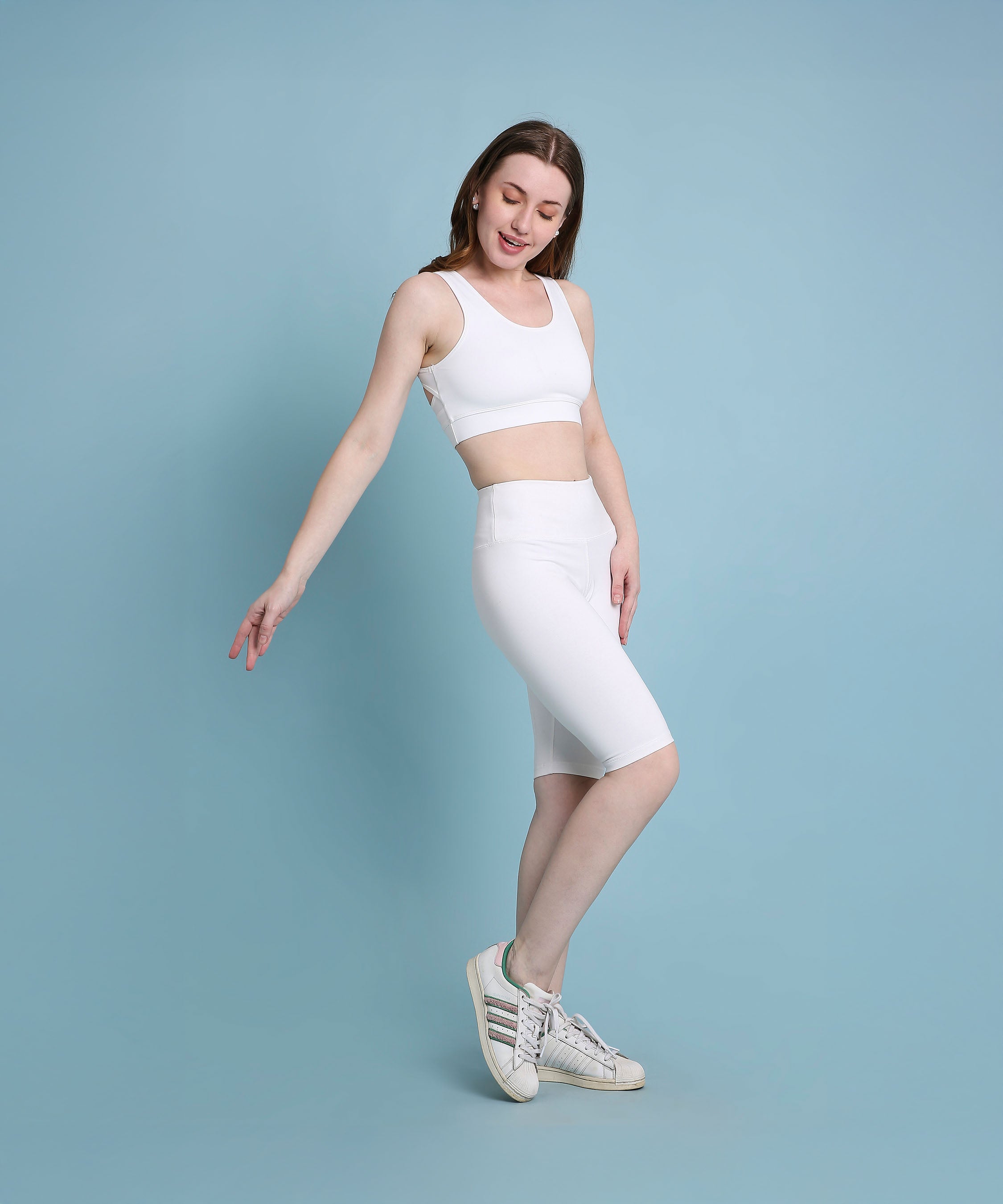 A woman models the White Sports Bra, wearing white activewear and posing against a light blue background with a slight smile.