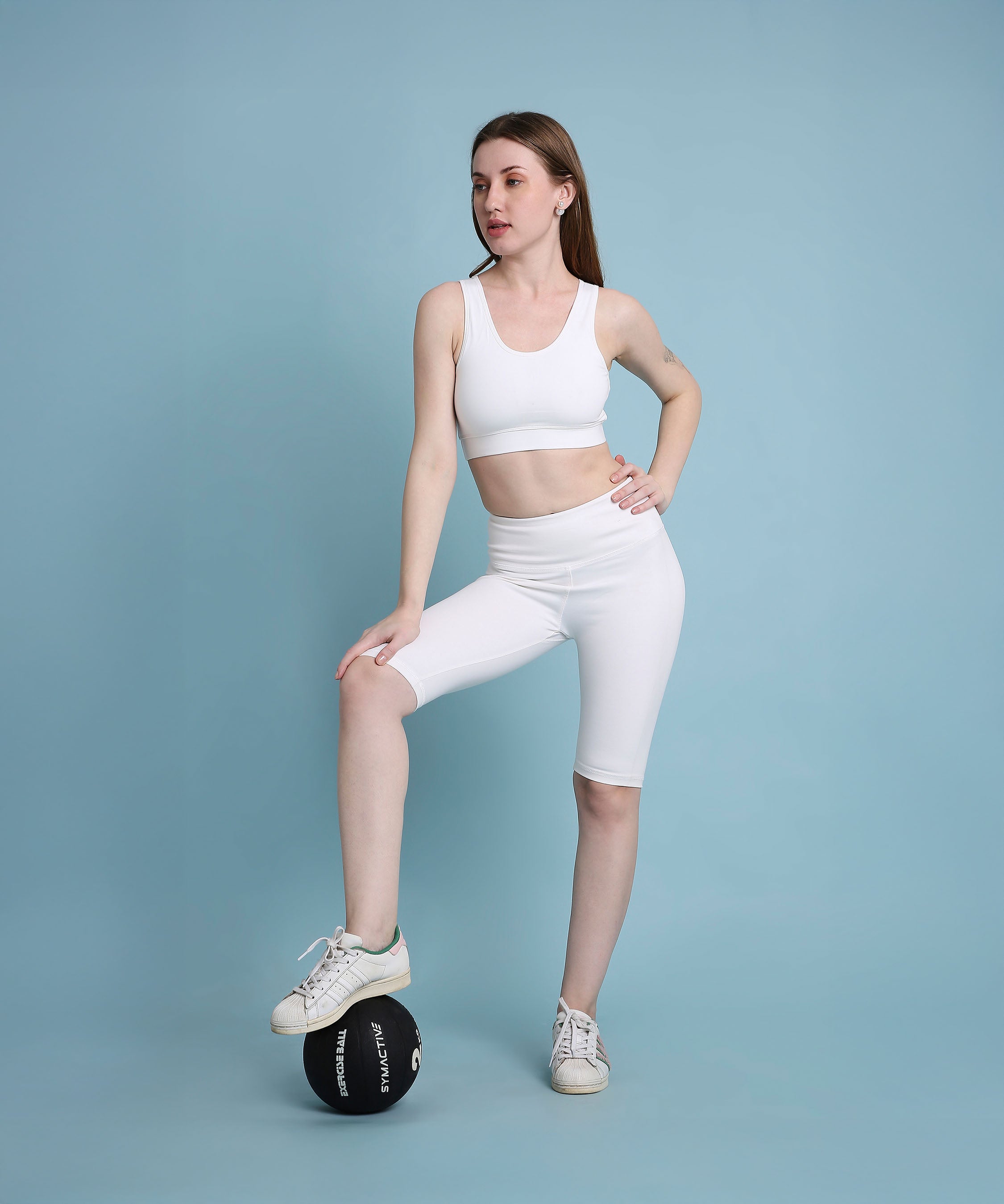 A woman in the White Sports Bra and athletic wear stands with one foot on a black medicine ball against a blue background, highlighting its medium support and moisture-wicking fabric.