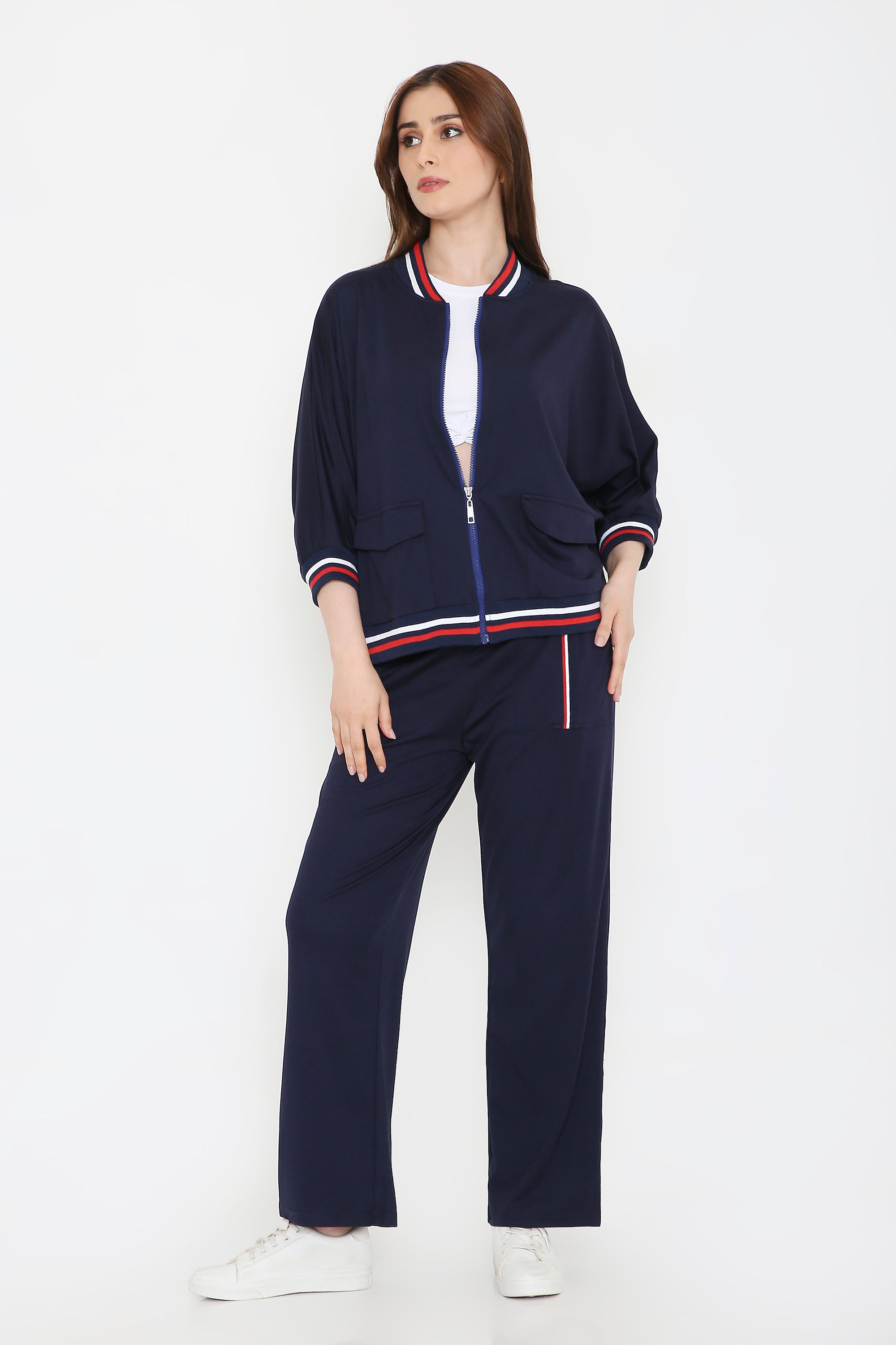 A woman in a Navy Blue Zipped Long Jacket with Tracks and white sneakers stands with one hand in her pocket against a white background.