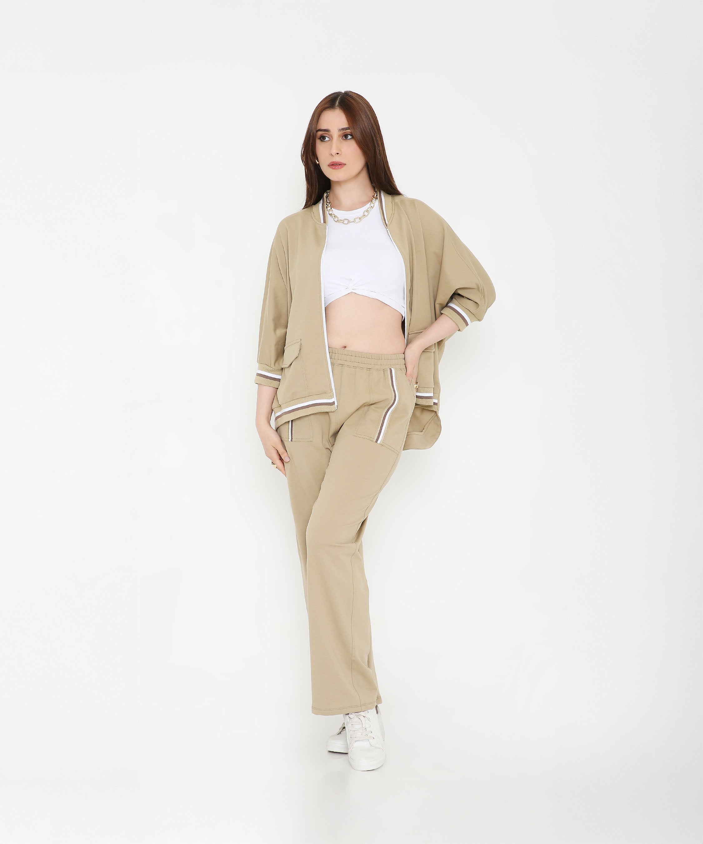 Woman in a Beige Zipped Long Jacket with Tracks and sneakers poses against a plain white background, showcasing chic style paired with the comfort of women’s activewear.