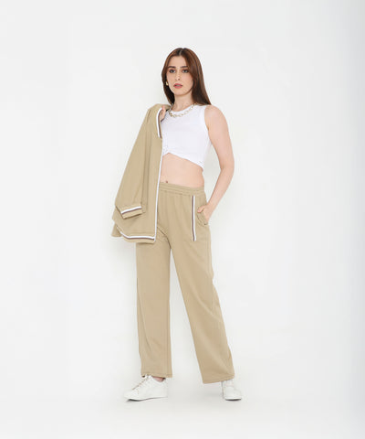 A woman in beige pants and white crop top holds the Beige Zipped Long Jacket with Tracks, standing against a white background—a modern, versatile track set for women.