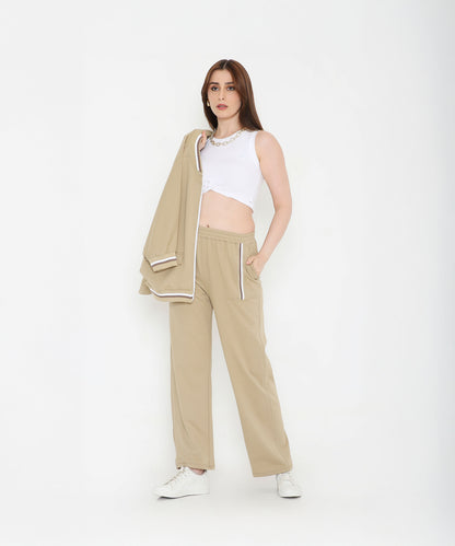 A woman in beige pants and white crop top holds the Beige Zipped Long Jacket with Tracks, standing against a white background—a modern, versatile track set for women.