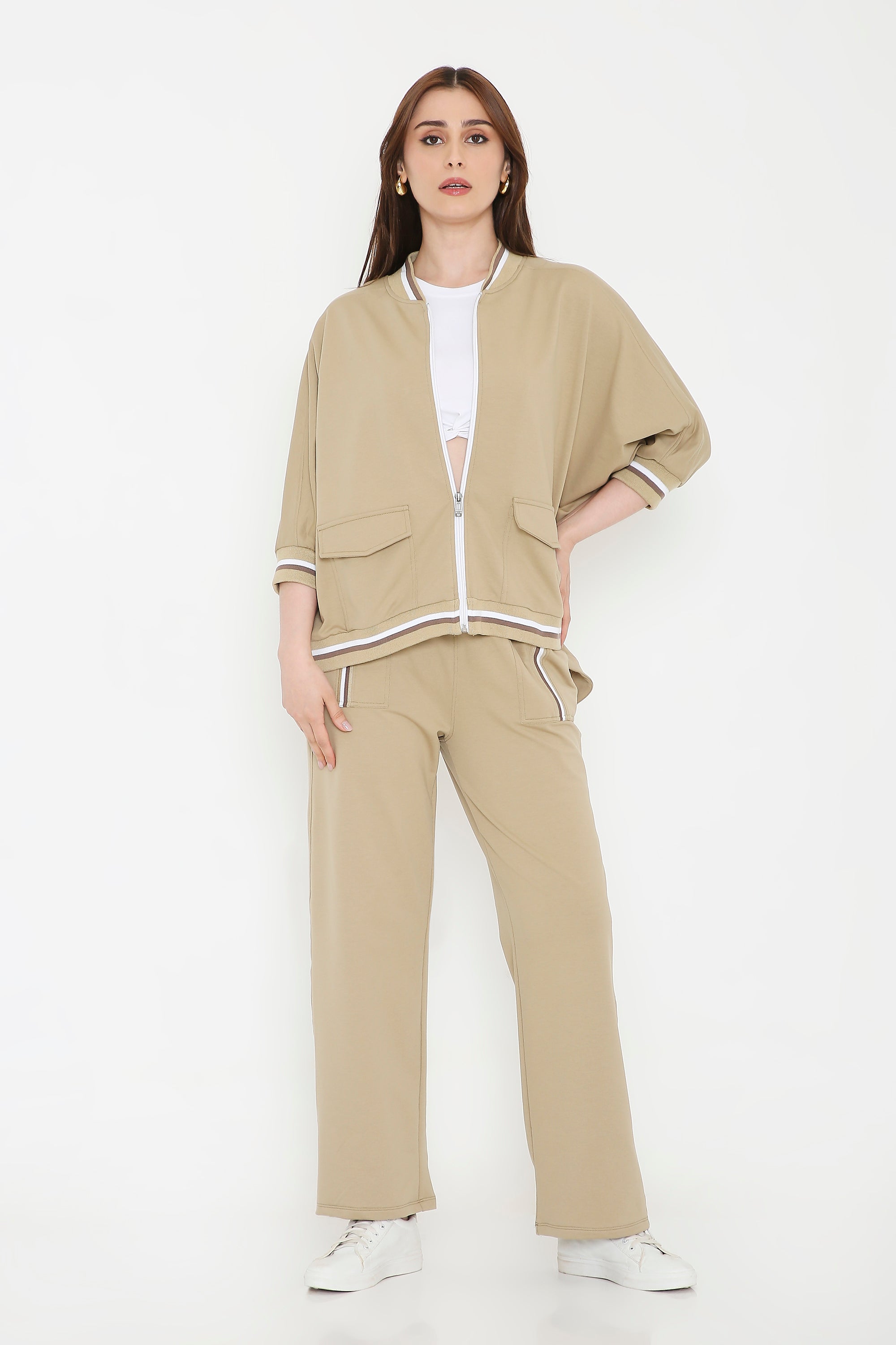 Woman wearing a Beige Zipped Long Jacket with Tracks, styled with a white crop top and sneakers, stands against a white background.