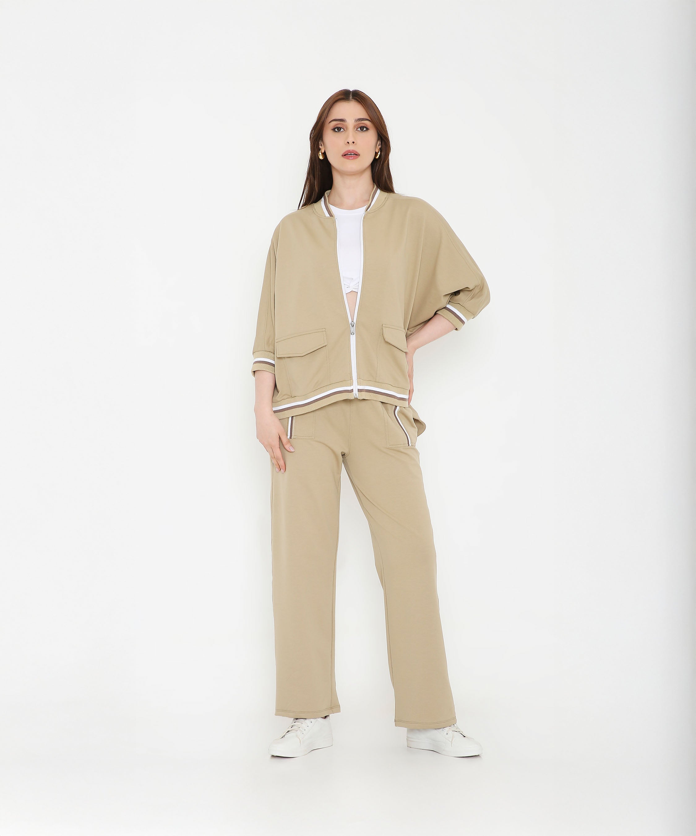 Woman wearing a Beige Zipped Long Jacket with Tracks, styled with a white crop top and sneakers, stands against a white background.