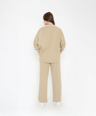 Woman facing away, dressed in the Beige Zipped Long Jacket with Tracks and white sneakers, standing against a white background.