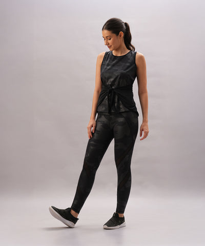 A woman wears the Metallic Tie-Up - Top Only in black camo with a shiny finish, standing on one leg and looking down, set against a neutral gray background.